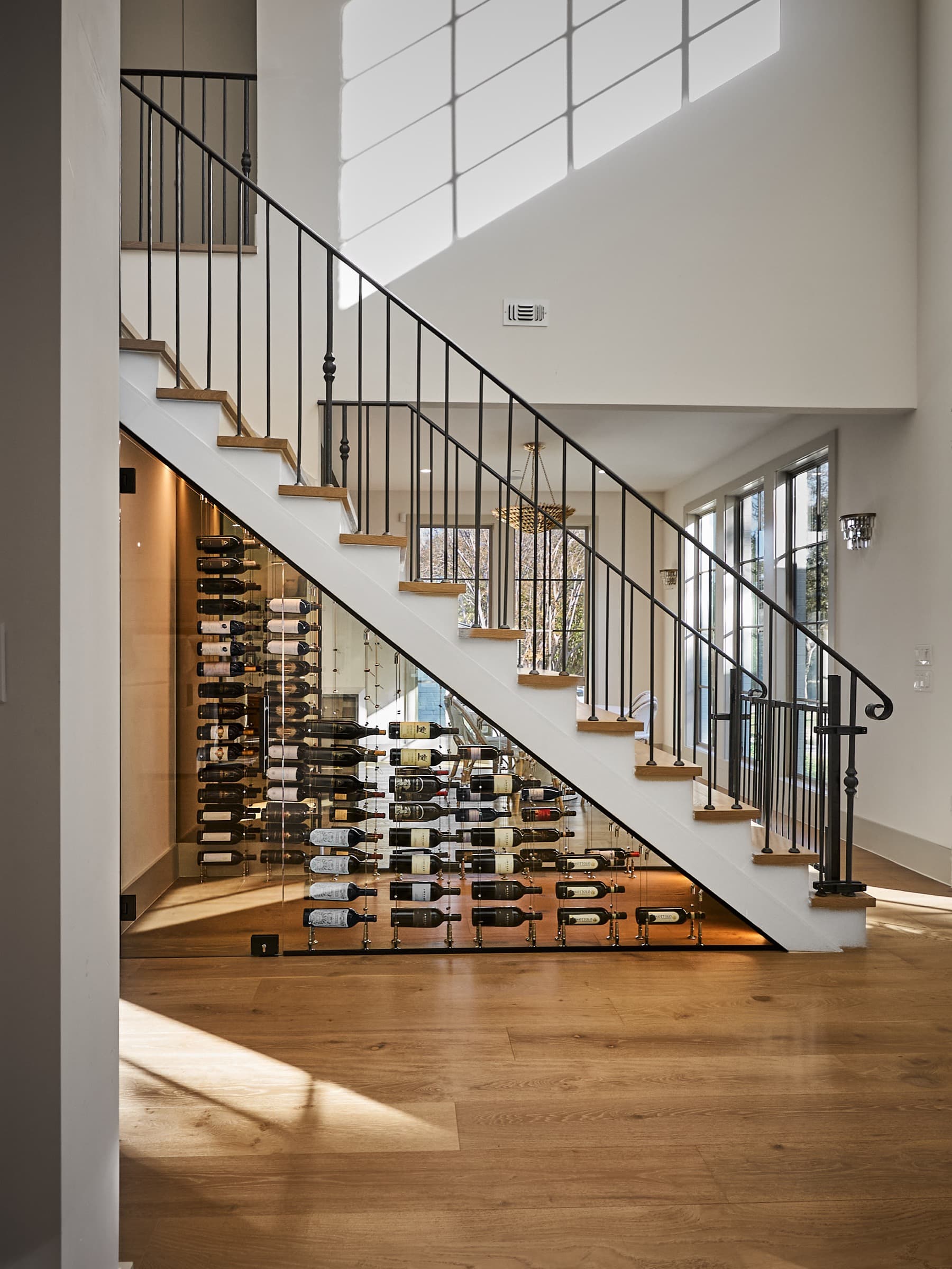 Completed passive under-stairs wine cellar in a West Austin luxury estate. Polished chrome cable racking behind Starphire glass, bottles loaded, natural light from the two-story stairwell windows, wide-plank oak hardwood floors