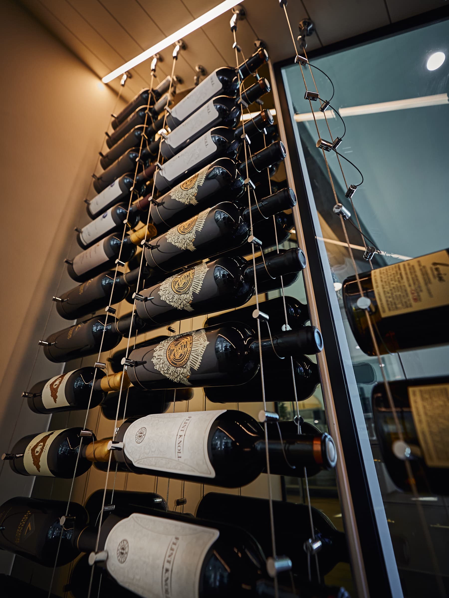 Dramatic low-angle view of wine bottles on polished chrome cable suspension racking inside a passive wine cellar. Recessed LED track lighting above, Starphire glass panel visible, ornate gold wine labels catching the light