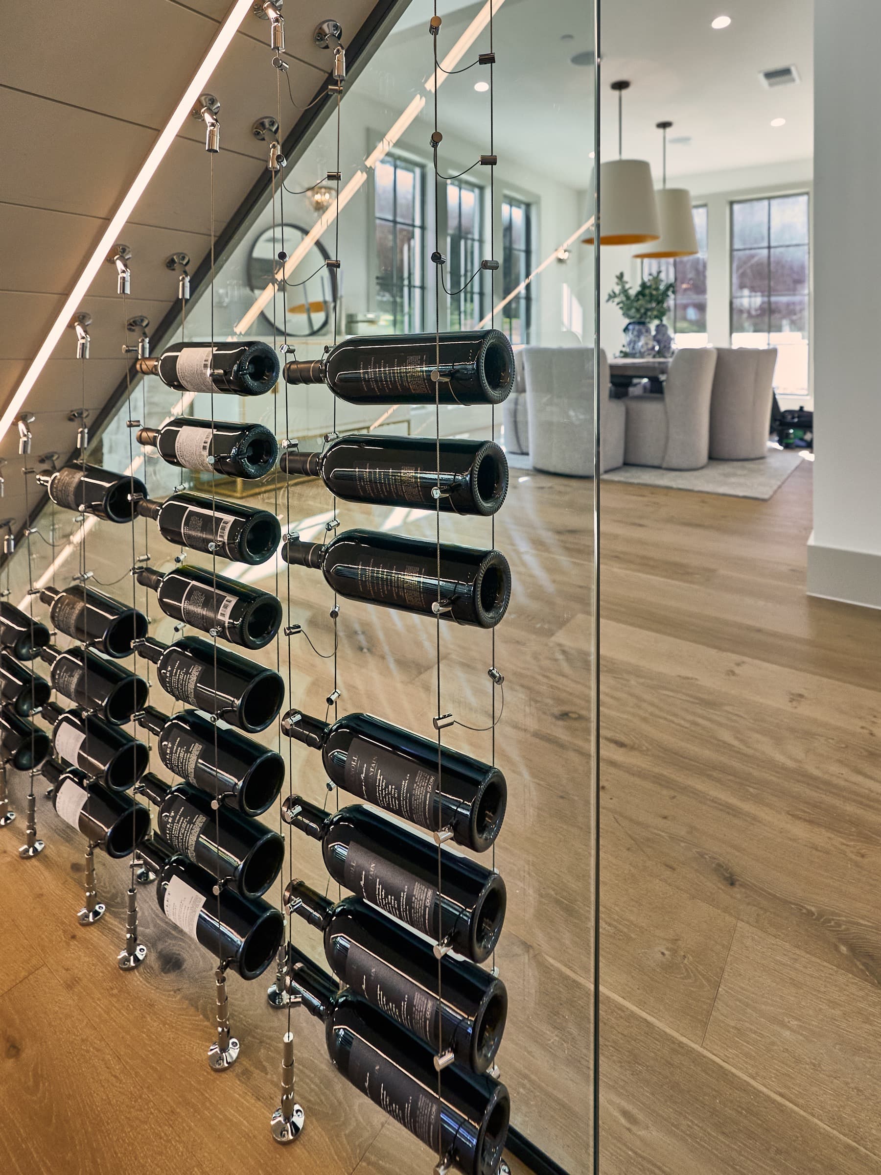 Interior view of the passive under-stairs wine cellar looking through frameless Starphire glass toward the living room. Bottles on polished chrome cable racks following the stair angle, LED accent strip visible, modern furniture and pendant lights in the background