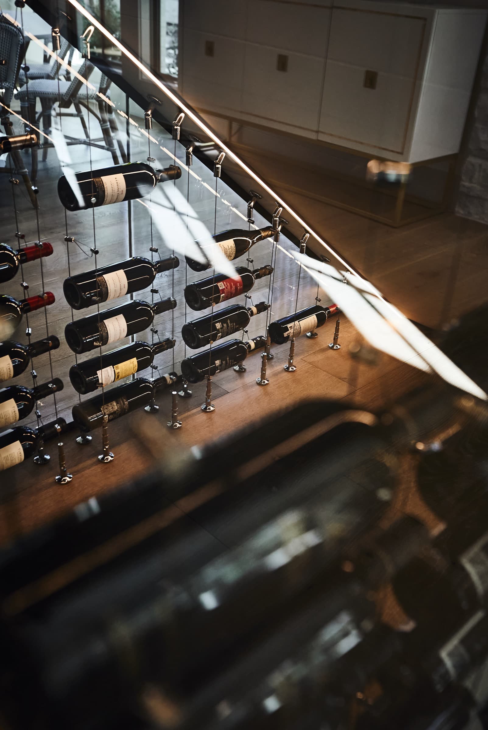 Overhead view looking down through a grand staircase at the under-stairs passive wine cellar below. Sunlight streaking across bottles on chrome cable racks, seen through Starphire glass from the second-floor landing