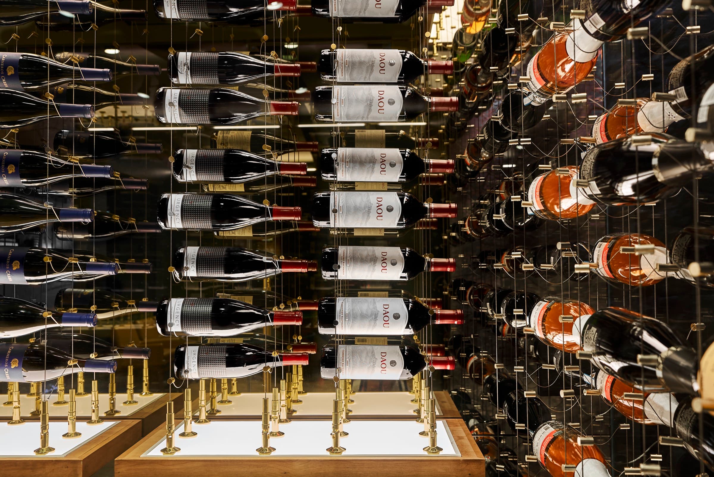 Close-up detail of the Bijou house wine cellar's cable-suspension display wall, showing label-forward bottles of DAOU Cabernet Sauvignon, Urgency, and Carneros Creek against a mirrored back wall