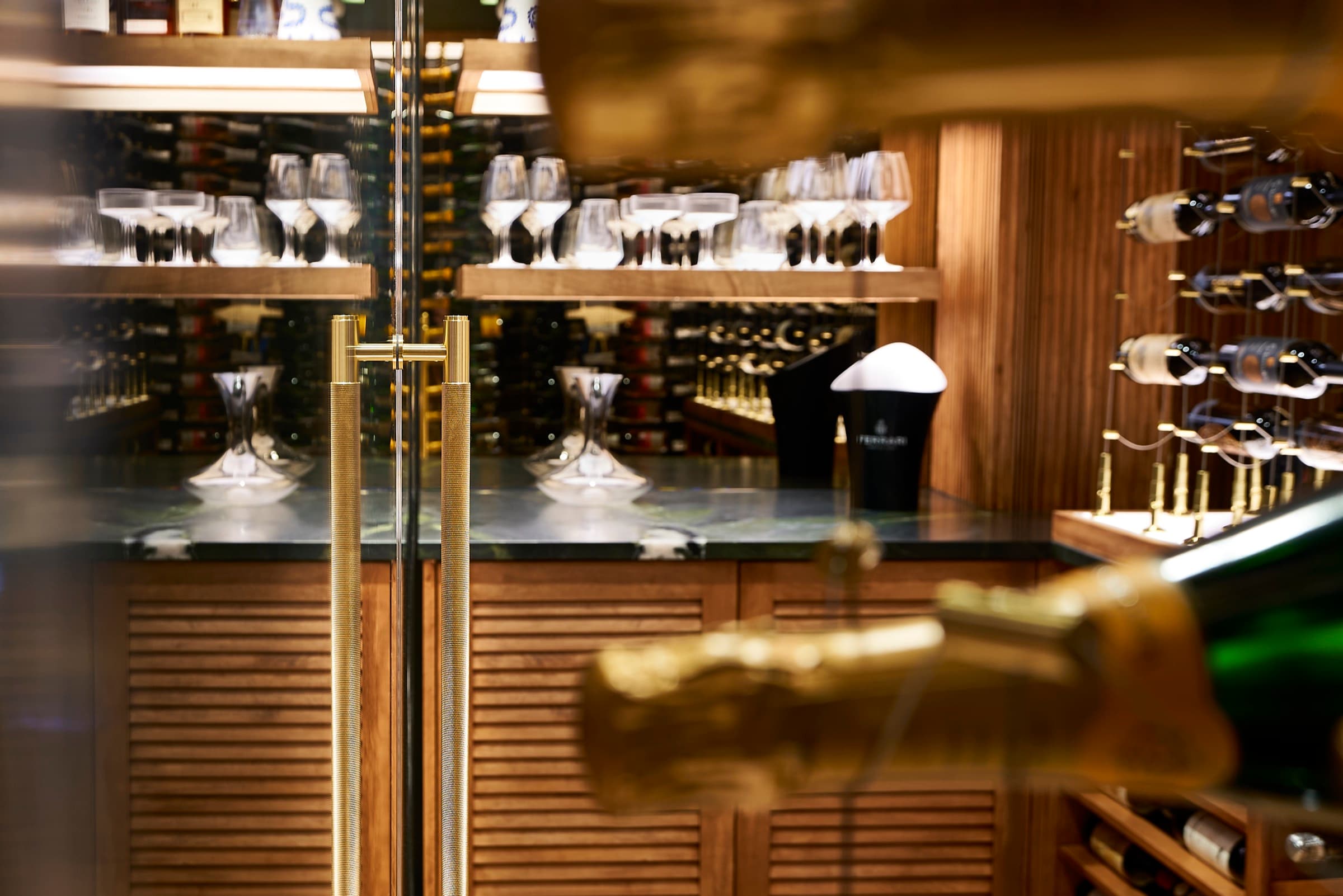 Through the frameless glass door of the Bijou house wine cellar, showing the interior tasting bar with brass T-bar door handles, two crystal decanters, a Ferrari Trento champagne bucket, and a backlit walnut stemware shelf